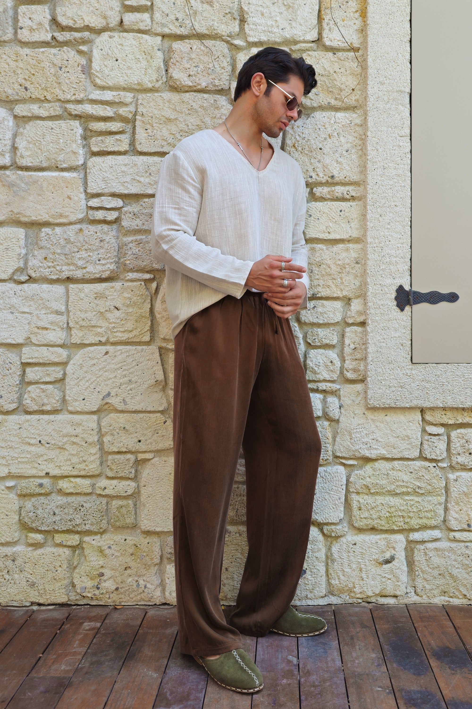 Man wearing a white sweater and brown pants standing against a stone wall.