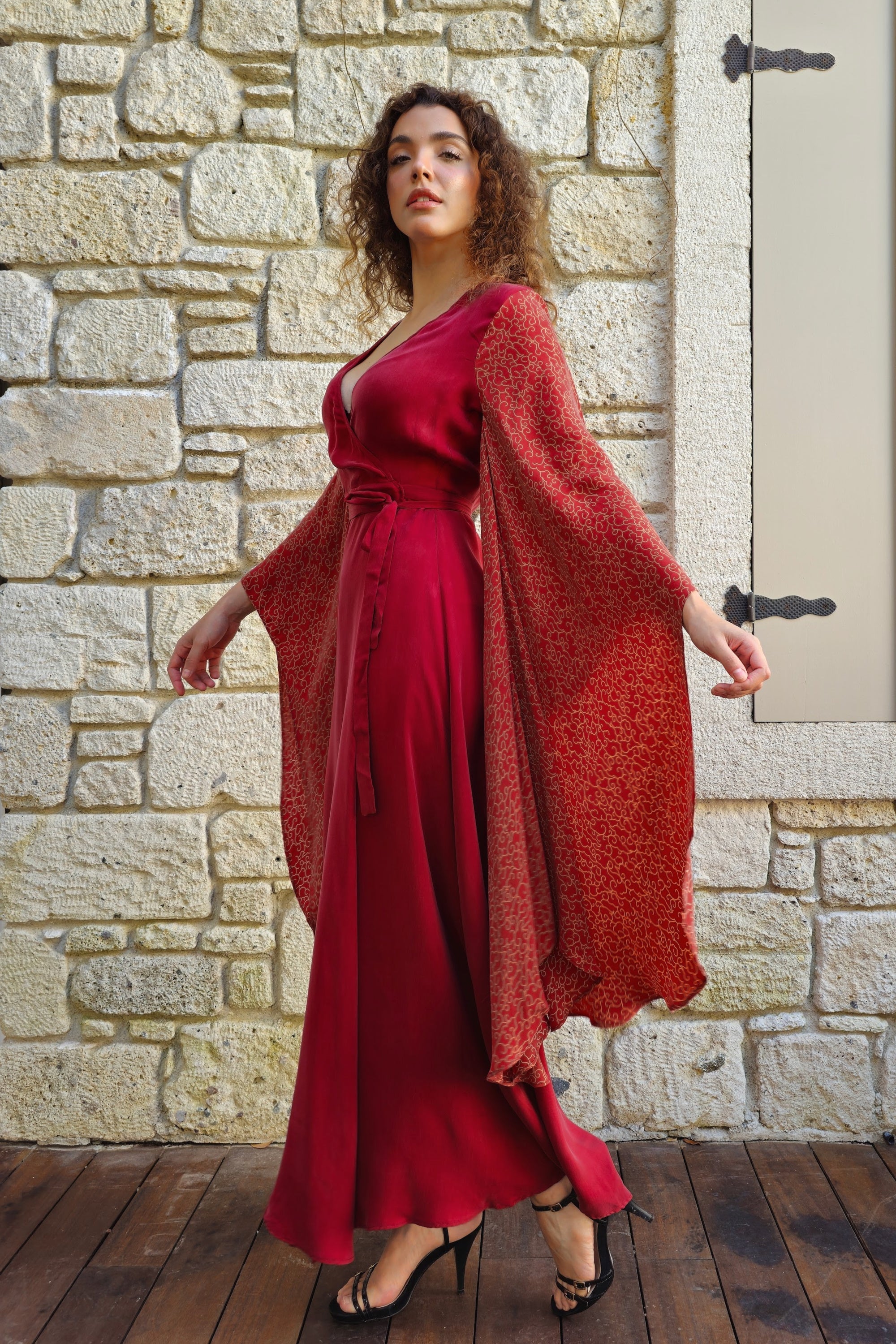Woman in a red dress and matching cardigan standing against a stone wall.