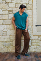 Man wearing a teal sleeveless shirt and brown pants standing against a stone wall.