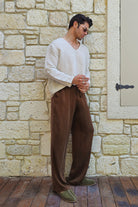 Man wearing a white sweater and brown pants standing against a stone wall.