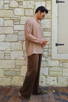 Man in beige shirt and brown pants standing against a stone wall.
