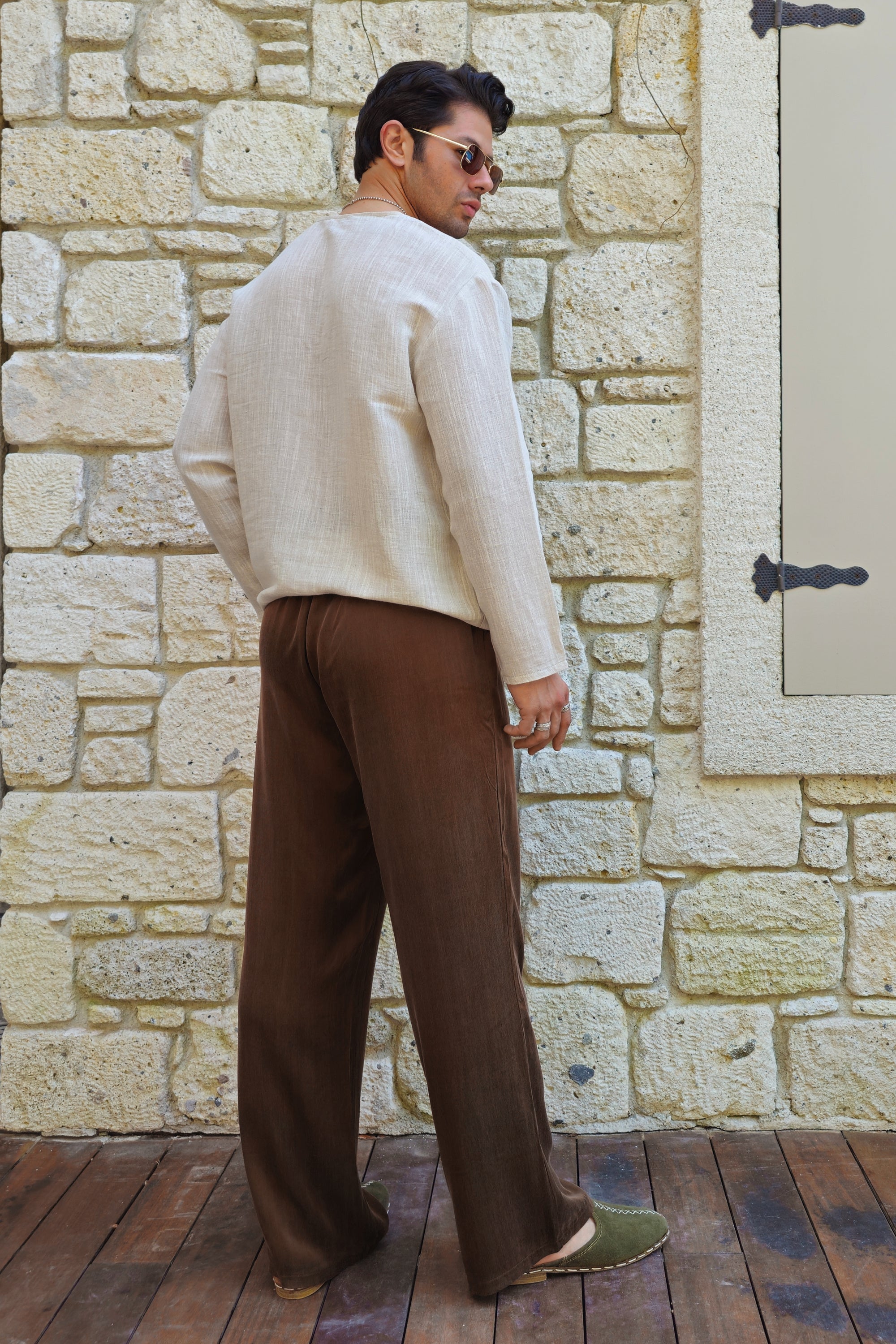 Man wearing a beige sweater and brown pants standing against a stone wall.